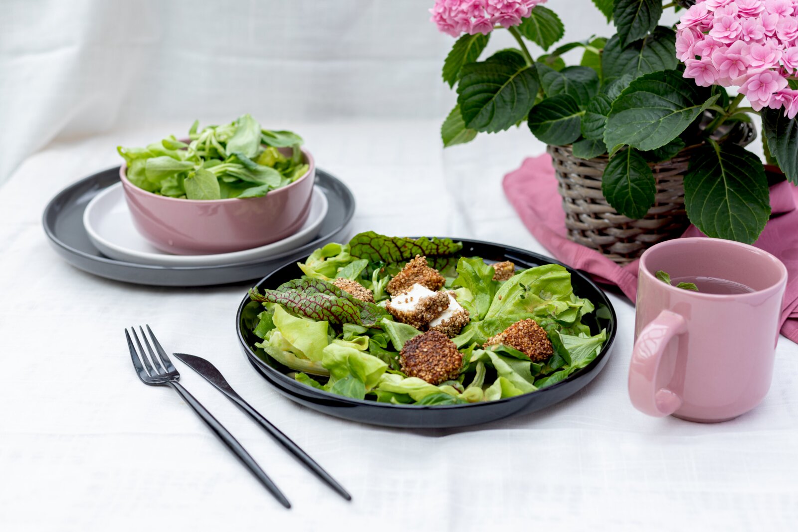 Schwarzer Teller mit frischem Blattsalat und panierten Käsewürfeln auf weißer Tischdecke, daneben rosa Tasse und Schüssel sowie rosa Hortensie im Korb – Perspektive: leicht von vorne in schräger Draufsicht.