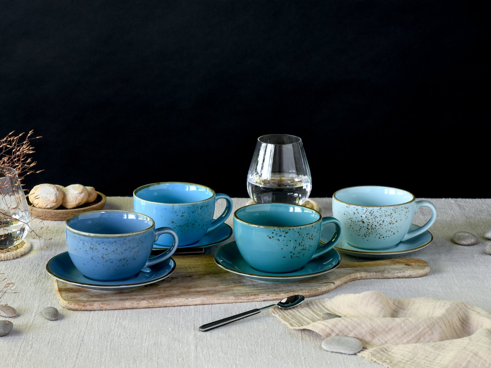 Vier blau-türkise Keramik-Kaffeetassen mit passenden Untertassen und Sprenkelglasur auf Holzbrett und Tischdecke, mit Wasserglas im Hintergrund; perspektive: frontale Ansicht auf Tischhöhe, leicht von oben