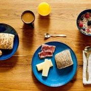 Draufsicht auf einen Frühstückstisch aus Holz mit blauem Teller mit Käse, Schinken und Mohnbrötchen, daneben Kaffee, Orangensaft, Müsli mit Beeren sowie Löffel und Besteck.