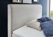 Seitliche Ansicht eines Boxspringbetts mit gepolstertem Kopfteil in hellem Stoff, dekoriert mit blauen und beigen Kissen.