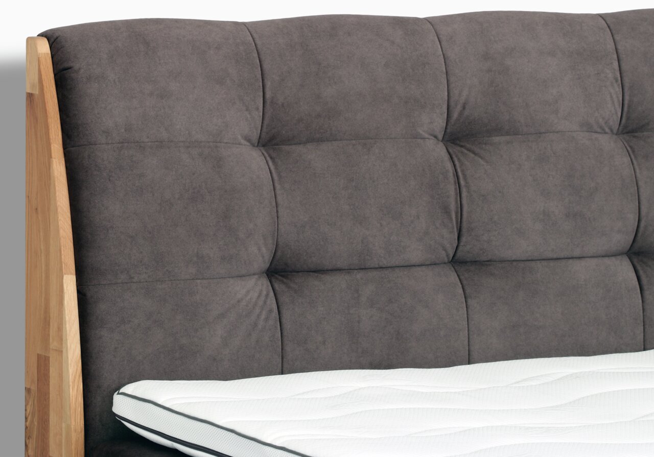 Nahaufnahme eines Boxspringbettes mit gepolstertem, grauem Kopfteil und seitlichem Holzrahmen, von der Seite betrachtet.