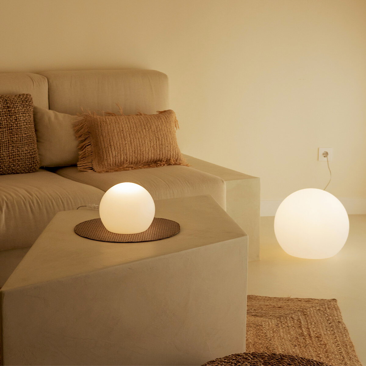 Minimalistisches Wohnzimmer mit beigem Sofa und Kissen; zwei leuchtende, kugelförmige Boden-/Tischlampen in warmweiß, eine auf dem Sofaelement und eine am Boden neben der Wandsteckdose, aufgenommen aus leicht erhöhter, schräger Frontperspektive.
