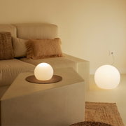 Minimalistische woonkamer met een beige bank en kussens; twee heldere, bolvormige staande lampen of tafellampen met warm wit licht, één op de bank en één op de vloer naast het stopcontact, gefotografeerd vanuit een licht verhoogd, schuin vooraanzicht.