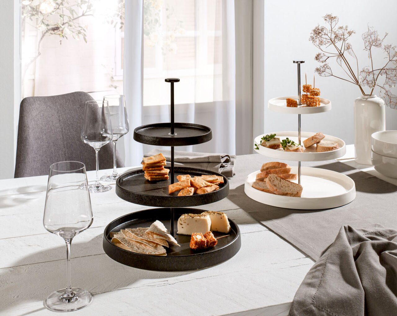 Zwei Etageren aus Porzellan, eine in Schwarz und eine in Weiß, auf einem gedeckten Tisch mit Snacks und Weingläsern, seitliche Perspektive.