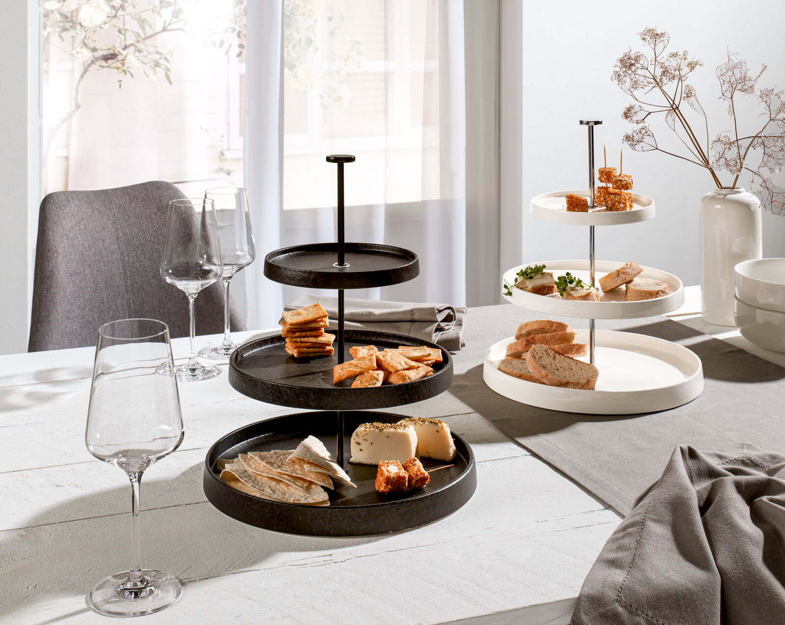 Zwei Etageren aus Porzellan, eine in Schwarz und eine in Weiß, auf einem gedeckten Tisch mit Snacks und Weingläsern, seitliche Perspektive.