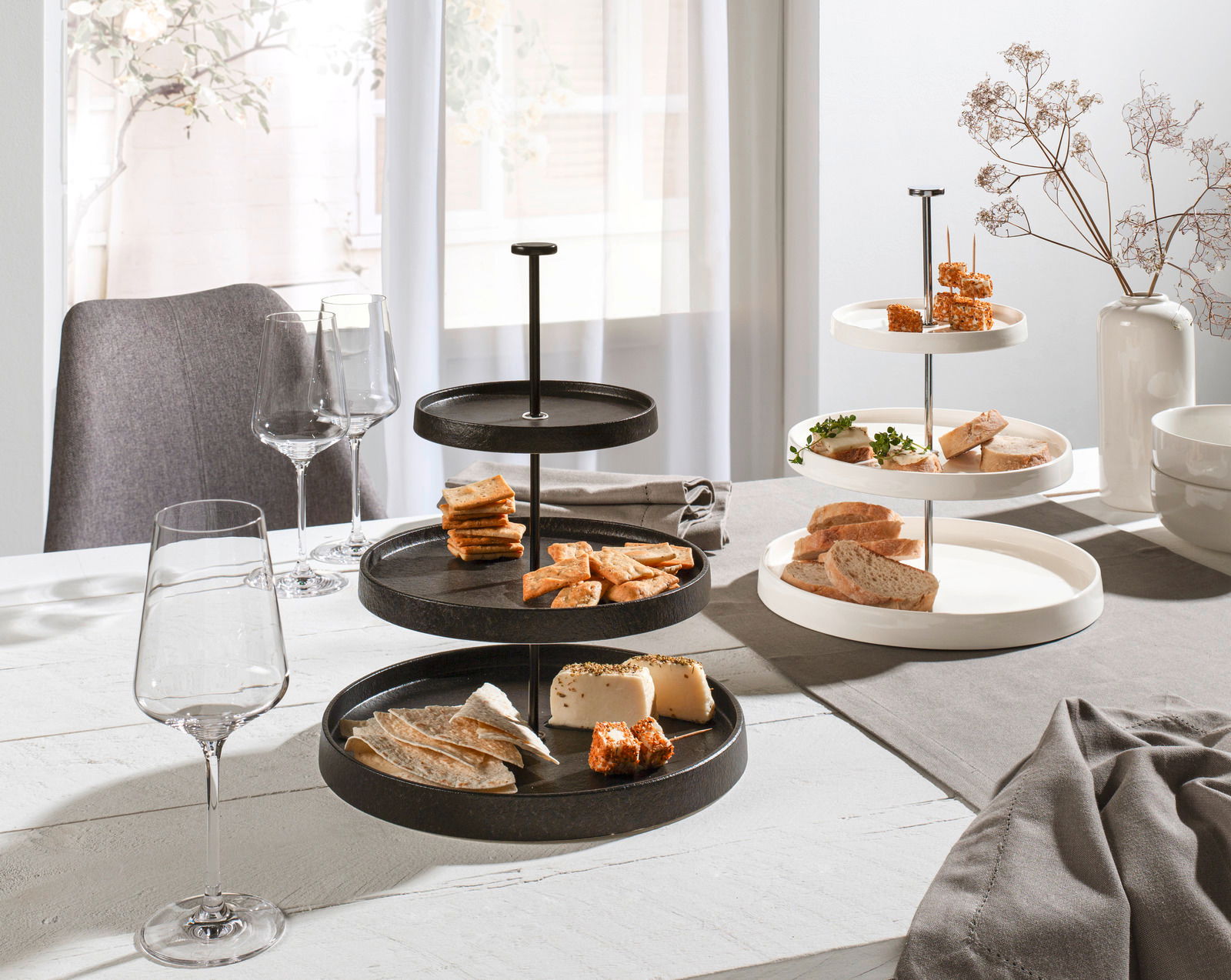 Zwei Etageren aus Porzellan, eine in Schwarz und eine in Weiß, auf einem gedeckten Tisch mit Snacks und Weingläsern, seitliche Perspektive.