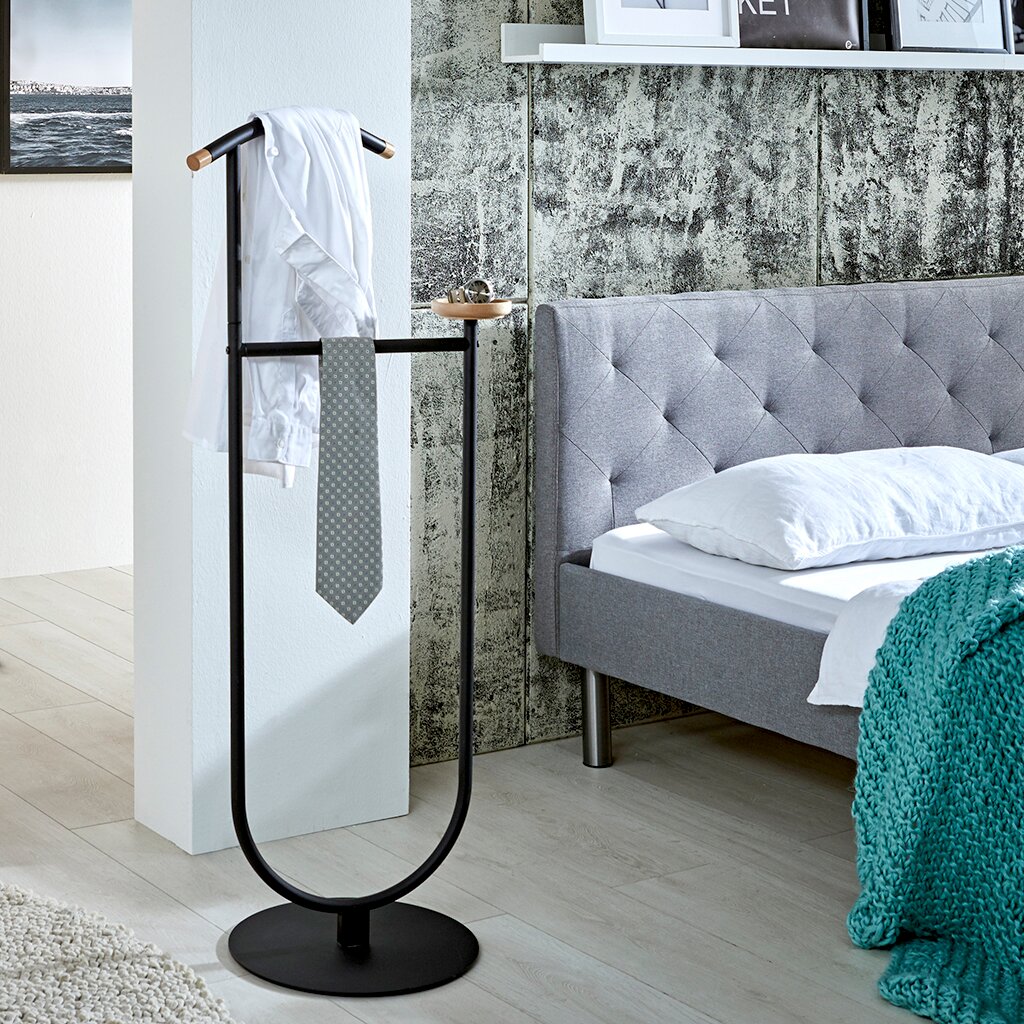 Schwarzer, freistehender Kleiderdiener aus Metall mit Holzdetails, Hemd und Krawatte aufgehängt, seitliche Perspektive im Schlafzimmer neben grauem Bett.