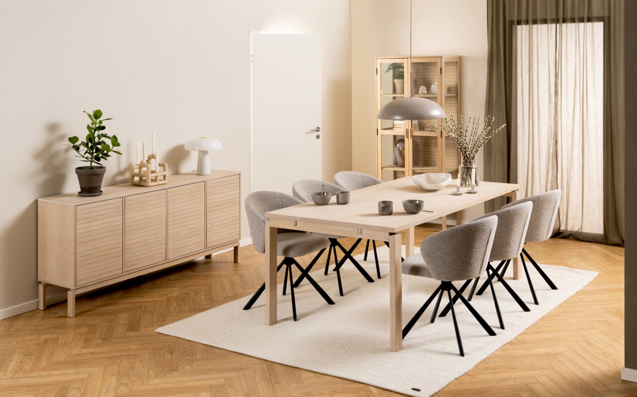 Modernes Sideboard aus hellem Holz mit vier Türen, dekoriert mit einer Pflanze, Kerzenhalter und Lampe, seitliche Perspektive.