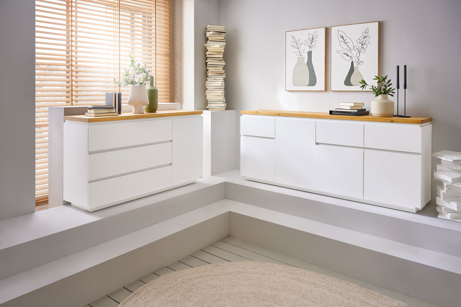 Novel XL-dressoir DALTON Zwei moderne, weiße Sideboards mit Holzoberfläche in einem hellen Raum, seitliche Perspektive.