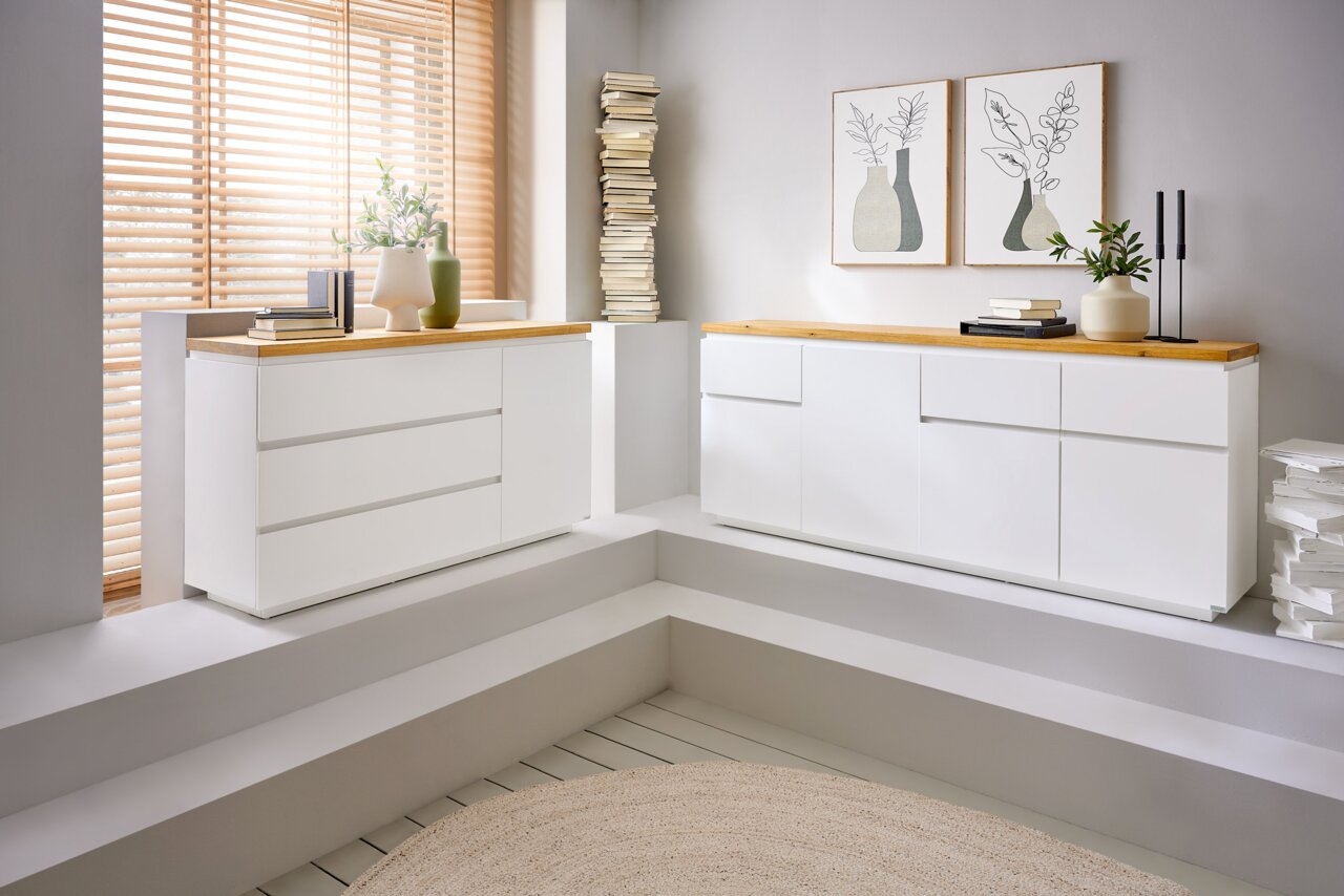 Novel Sideboard DALTON Zwei moderne, weiße Sideboards mit Holzoberfläche in einem hellen Raum, seitliche Perspektive.