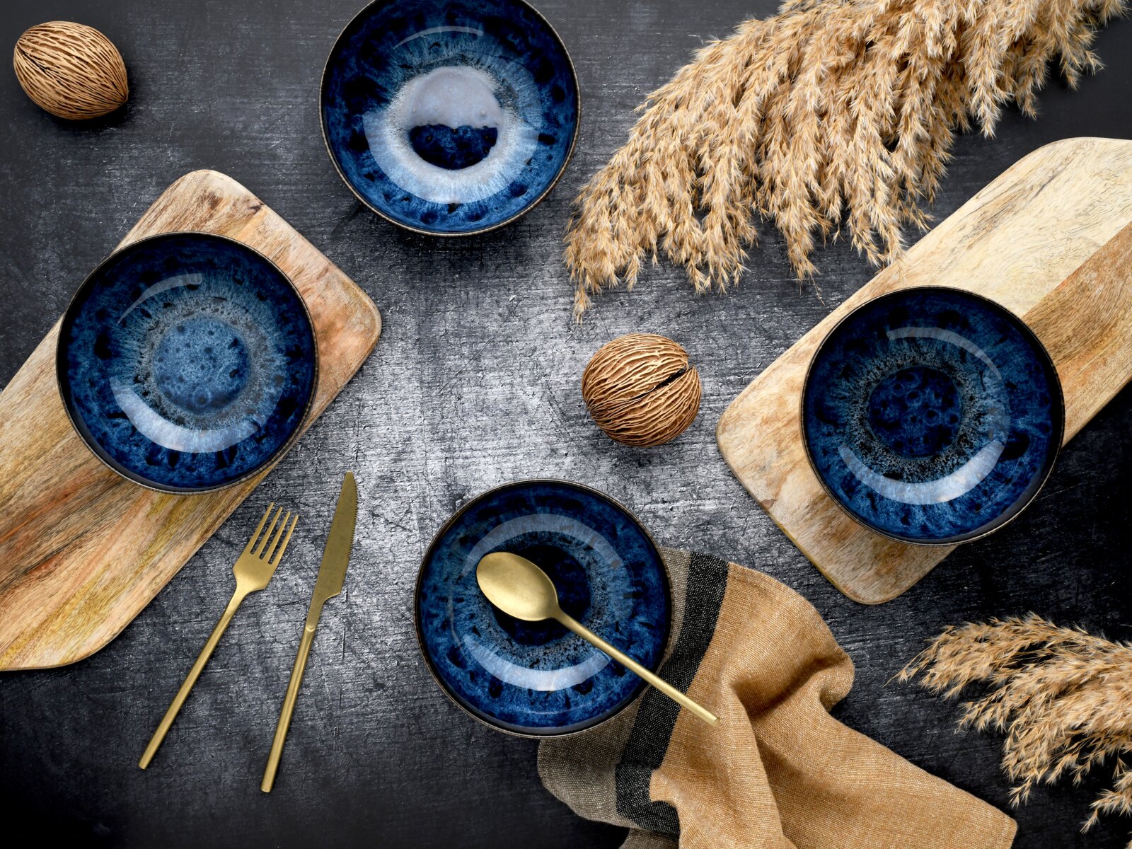 Vier tiefblaue Keramikschalen mit reaktiver Glasur auf Holzbrettchen auf dunklem Tisch, daneben goldenes Besteck, Deko-Nüsse und Trockengras; Draufsicht (Top-Down).