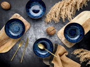 Vier tiefblaue Keramikschalen mit reaktiver Glasur auf Holzbrettchen auf dunklem Tisch, daneben goldenes Besteck, Deko-Nüsse und Trockengras; Draufsicht (Top-Down).