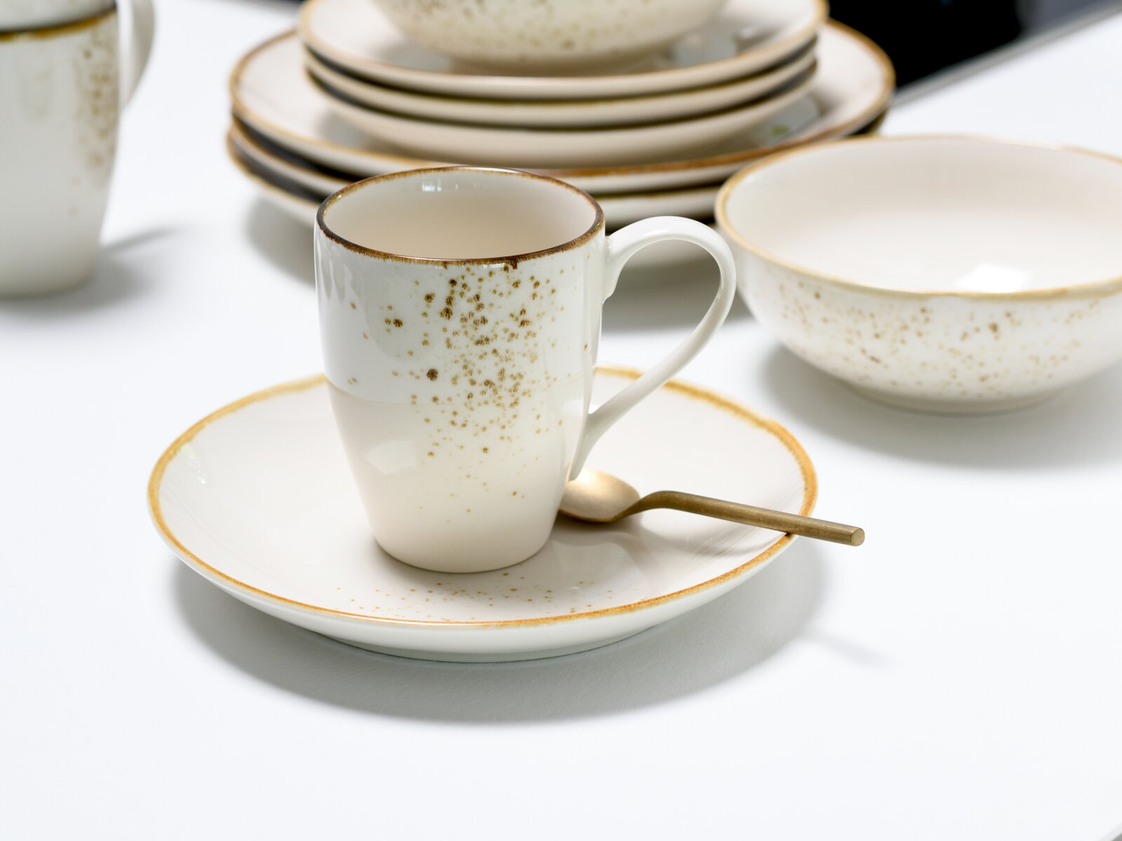 Gesprenkelte weiße Porzellan-Kaffeetasse mit Henkel auf passender Untertasse mit goldfarbenem Rand, seitliche Ansicht in leicht erhöhter Perspektive; im Hintergrund Teller und Schale.