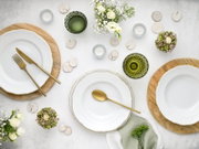 Draufsicht auf ein elegant gedecktes Tisch-Arrangement mit weißen Porzellantellern mit goldener Kante auf Holzuntertellern, goldfarbenem Besteck, grünen Kristallgläsern, Teelichtern und weißen Blumen auf hellem Hintergrund.