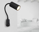 Schwarze Wandleuchte mit flexiblem Arm, seitliche Perspektive, montiert an einer weißen Wand neben einem modernen weißen Sofa.