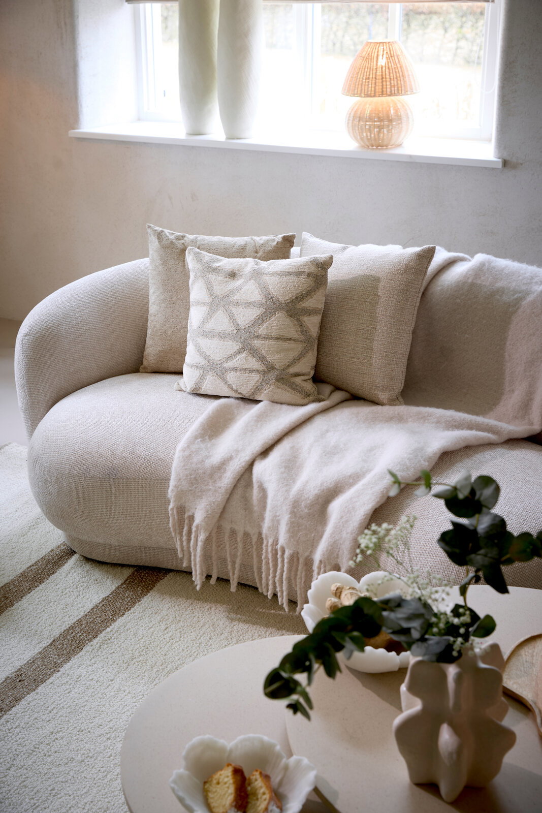 Gemütliches Wohnzimmer mit einer beigen Couch, dekoriert mit Kissen und einer Decke, im Vordergrund ein Couchtisch mit Pflanzen und Dekoration, seitliche Perspektive.