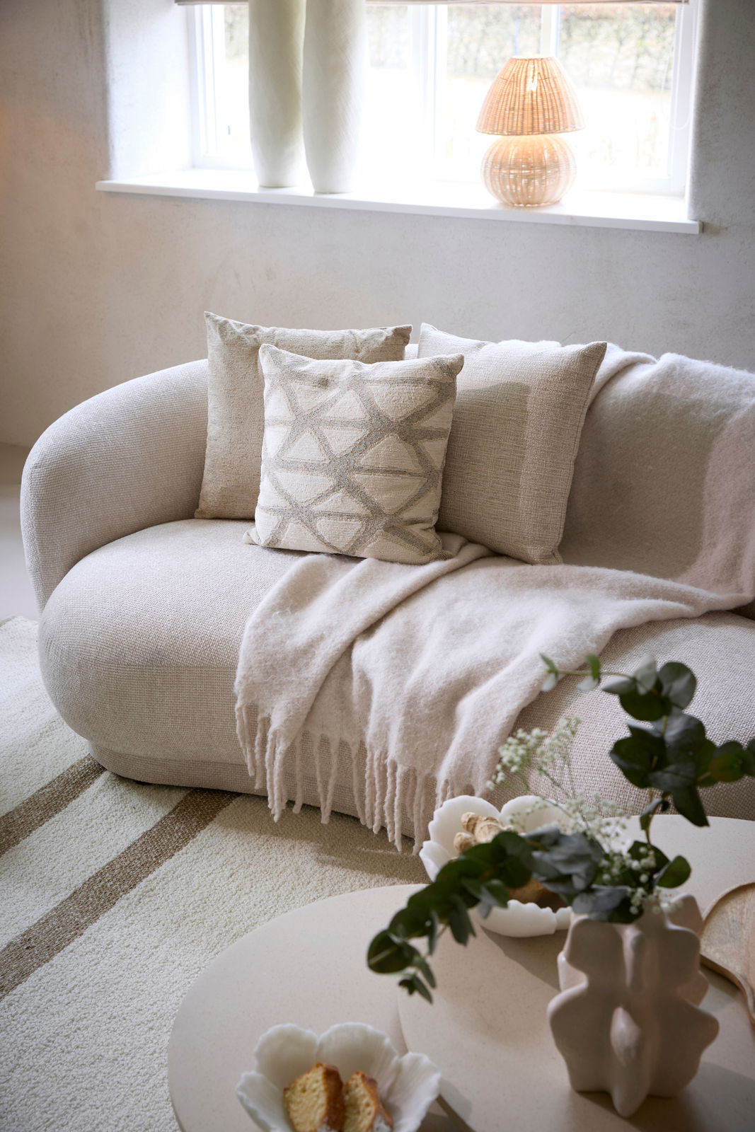 Gemütliches Wohnzimmer mit einer beigen Couch, dekoriert mit Kissen und einer Decke, im Vordergrund ein Couchtisch mit Pflanzen und Dekoration, seitliche Perspektive.