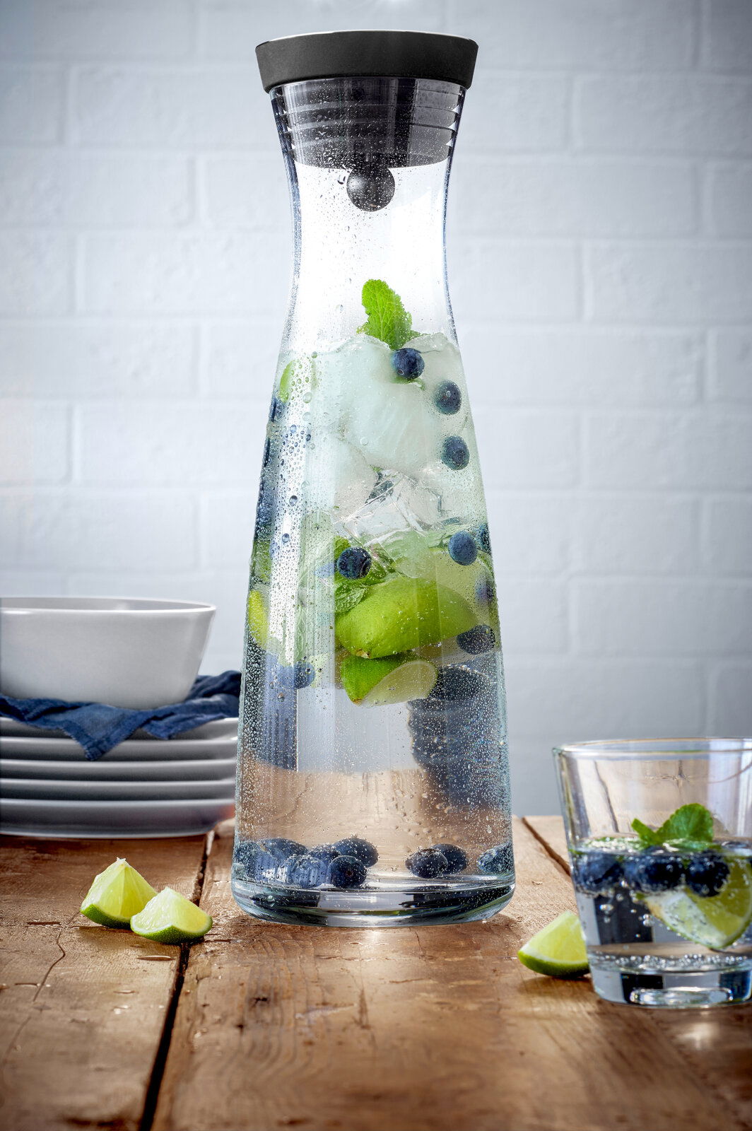 Wasserkaraffe BASIC 1,5L mit schwarzem Deckel, gefüllt mit Wasser, Eiswürfeln, Limettenscheiben, Blaubeeren und Minzblättern, auf einem Holztisch vor einer weißen Ziegelwand, seitliche Perspektive.