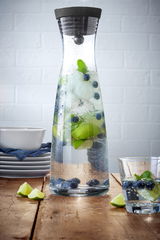 Wasserkaraffe BASIC 1,5L mit schwarzem Deckel, gefüllt mit Wasser, Eiswürfeln, Limettenscheiben, Blaubeeren und Minzblättern, auf einem Holztisch vor einer weißen Ziegelwand, seitliche Perspektive.