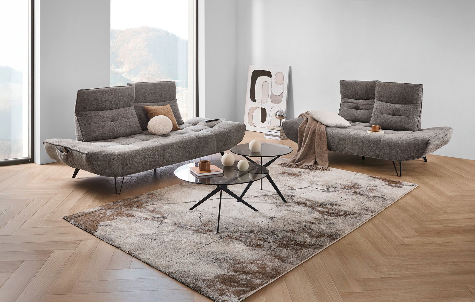 Moderne Polstergarnitur in einem hellen Raum mit großen Fenstern, bestehend aus zwei grauen Sofas mit verstellbaren Rückenlehnen, dekoriert mit Kissen und Decken, vor einem stilvollen Couchtisch auf einem gemusterten Teppich. Perspektive: Frontalansicht.
