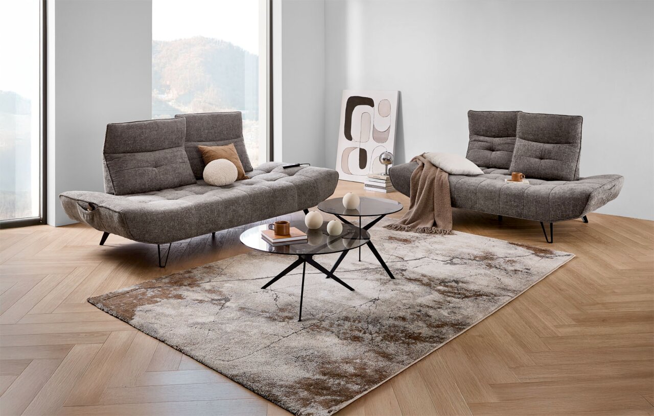 Moderne Polstergarnitur in einem hellen Raum mit großen Fenstern, bestehend aus zwei grauen Sofas mit verstellbaren Rückenlehnen, dekoriert mit Kissen und Decken, vor einem stilvollen Couchtisch auf einem gemusterten Teppich. Perspektive: Frontalansicht.