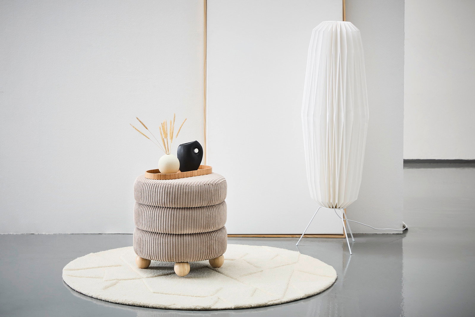 Moderner Hocker mit beiger, gerippter Polsterung und runden Holzfüßen, dekoriert mit einer Vase und einem Tablett, auf einem hellen Teppich in einem minimalistischen Raum. Seitliche Perspektive.