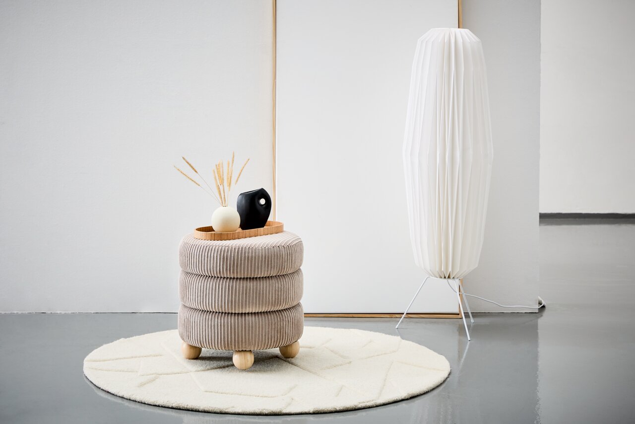 XORA Hocker MOLLY Moderner Hocker mit beiger, gerippter Polsterung und runden Holzfüßen, dekoriert mit einer Vase und einem Tablett, auf einem hellen Teppich in einem minimalistischen Raum. Seitliche Perspektive.