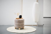 Moderner Hocker mit beiger, gerippter Polsterung und runden Holzfüßen, dekoriert mit einer Vase und einem Tablett, auf einem hellen Teppich in einem minimalistischen Raum. Seitliche Perspektive.