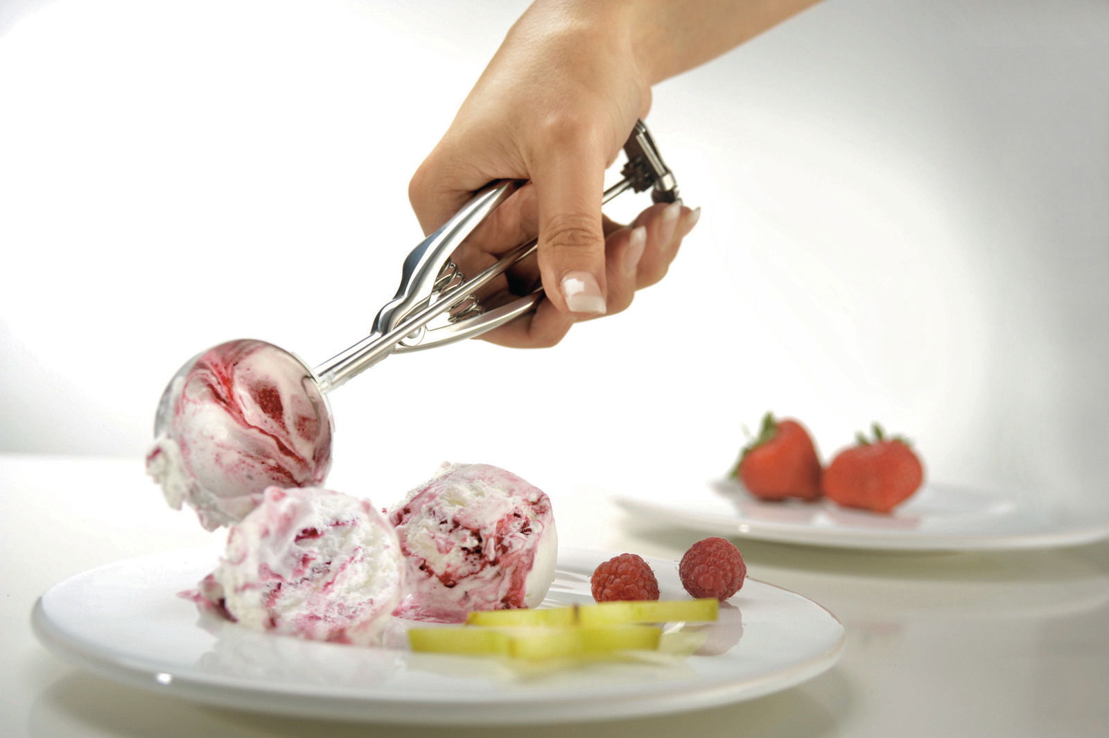 Nahaufnahme eines Eisportionierers, der eine Kugel Eiscreme serviert. Im Hintergrund sind frische Früchte auf einem Teller zu sehen.