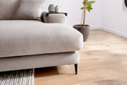Nahaufnahme einer modernen Longchairkombination in hellem Beige mit einem Kissen und einem kleinen Beistelltisch auf der Armlehne, auf dem eine Tasse und eine Teekanne stehen. Im Hintergrund ist ein Holzboden und eine Pflanze zu sehen.