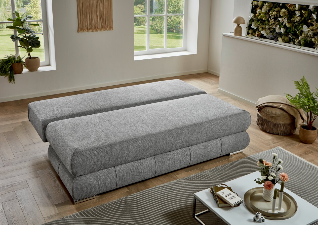 Graues Funktionssofa in flacher Position, seitliche Perspektive, in modernem Wohnzimmer mit Pflanzen und Dekoration.