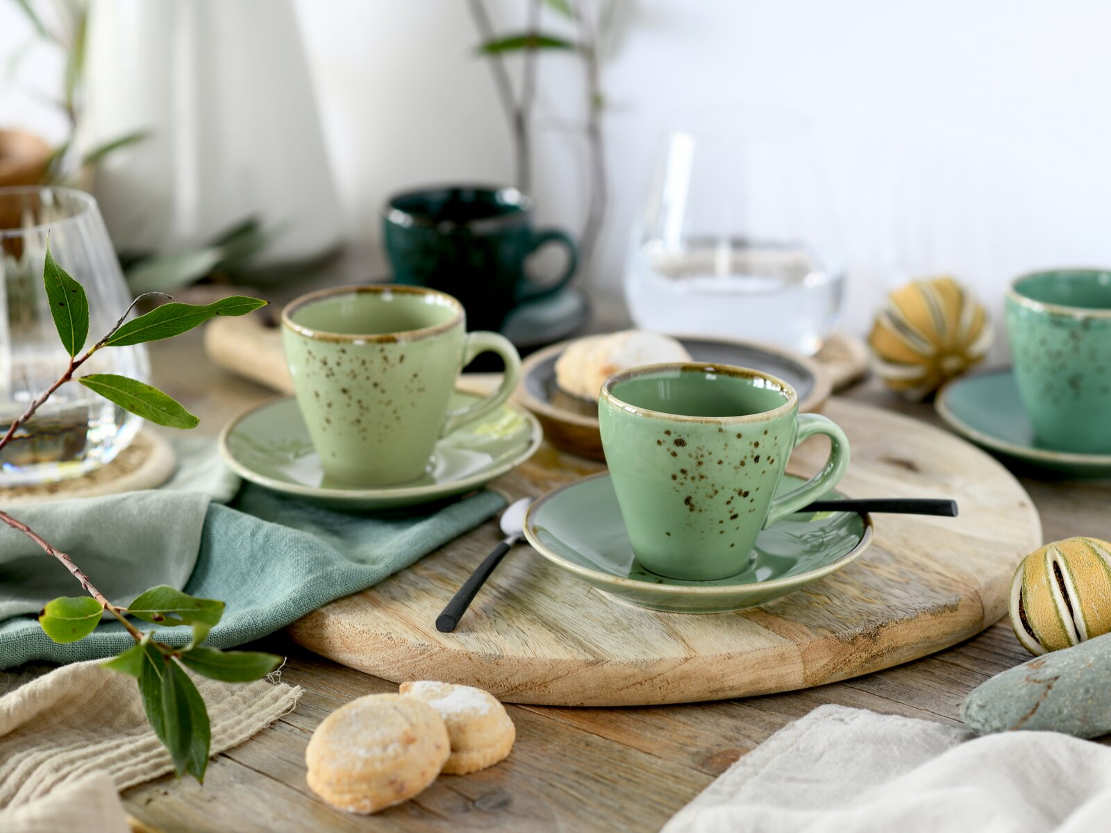 Zwei gesprenkelte, hellgrüne Keramik-Kaffeetassen mit Untertassen auf einem runden Holzbrett, mit Besteck und Gebäck auf rustikalem Tisch, fotografiert in leichter Schrägansicht von vorne.