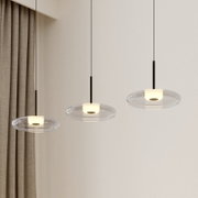 Drei moderne Pendelleuchten mit flachem, transparentem Glasschirm und warmweißem Licht an schwarzen Kabeln, Innenraumaufnahme in leichter Untersicht (Blick von unten schräg nach oben) vor heller Wand und beigem Vorhang.