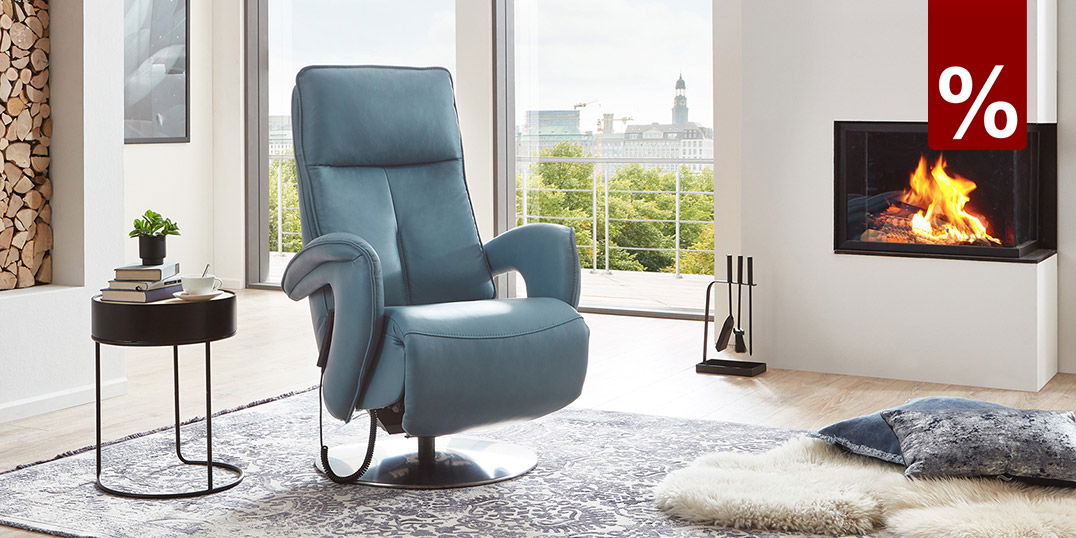 Moderner Wohnraum mit blauem Relaxsessel, Kaminfeuer und heller Einrichtung mit Holz und großen Fenstern.