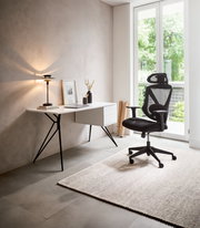 Moderner Schreibtisch mit schlanken schwarzen Beinen und weißer Tischplatte, seitlich mit Schubladen, in einem hellen Raum mit Blick auf einen ergonomischen Bürostuhl und ein großes Fenster.