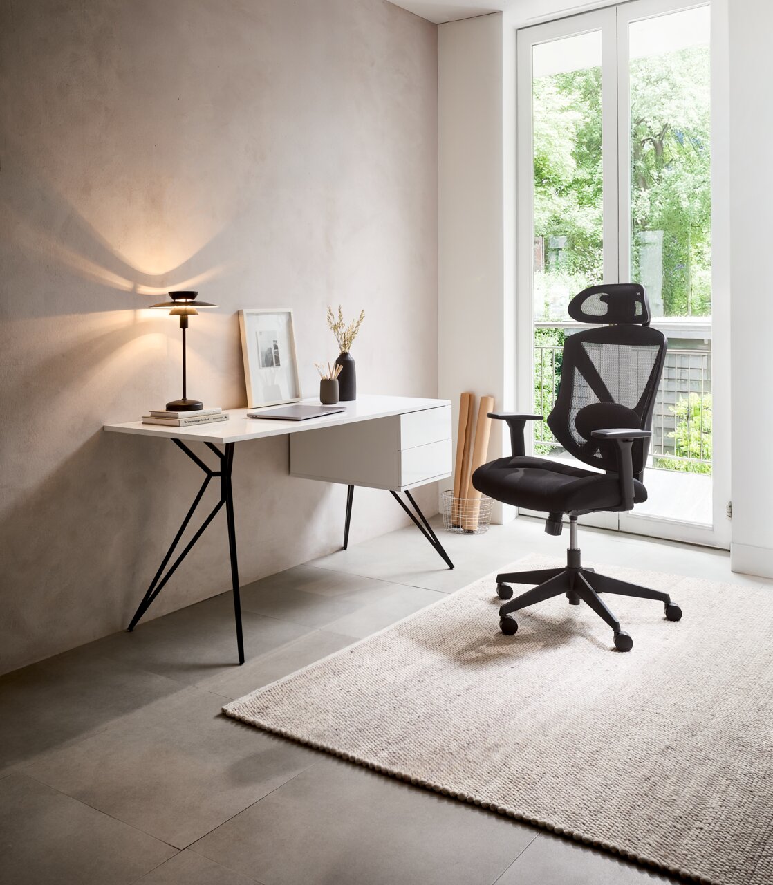 Moderner Schreibtisch mit schlanken schwarzen Beinen und weißer Tischplatte, seitlich mit Schubladen, in einem hellen Raum mit Blick auf einen ergonomischen Bürostuhl und ein großes Fenster.