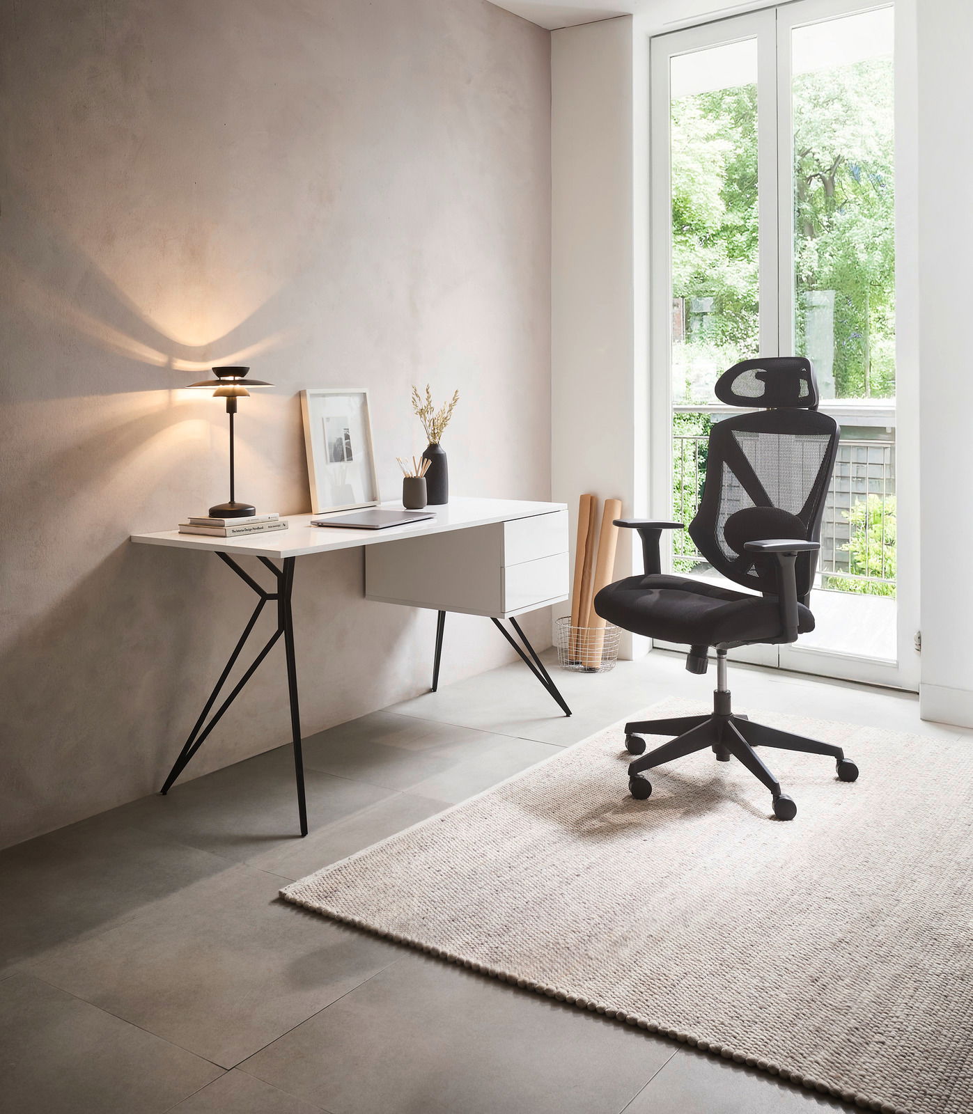 Moderner Schreibtisch mit schlanken schwarzen Beinen und weißer Tischplatte, seitlich mit Schubladen, in einem hellen Raum mit Blick auf einen ergonomischen Bürostuhl und ein großes Fenster.