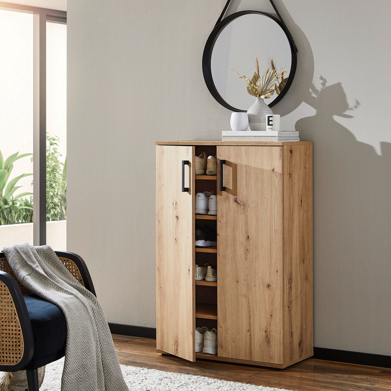 Schuhschrank aus Holz mit zwei Türen, teilweise geöffnet, zeigt mehrere Paar Schuhe. Der Schrank steht in einem modernen Raum mit einem runden Spiegel darüber. Perspektive: seitlich von vorne.