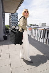 Frau mit Sonnenbrille trägt einen schwarzen Carrycruiser mit glänzenden Punkten auf einer Terrasse mit städtischem Hintergrund, seitliche Perspektive.