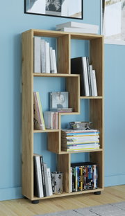 Modernes, asymmetrisches Bücherregal aus hellem Holz mit offenen Fächern, gefüllt mit Büchern, Zeitschriften und Deko; Aufnahme in leichter Schrägansicht von vorn vor blauer Wand.