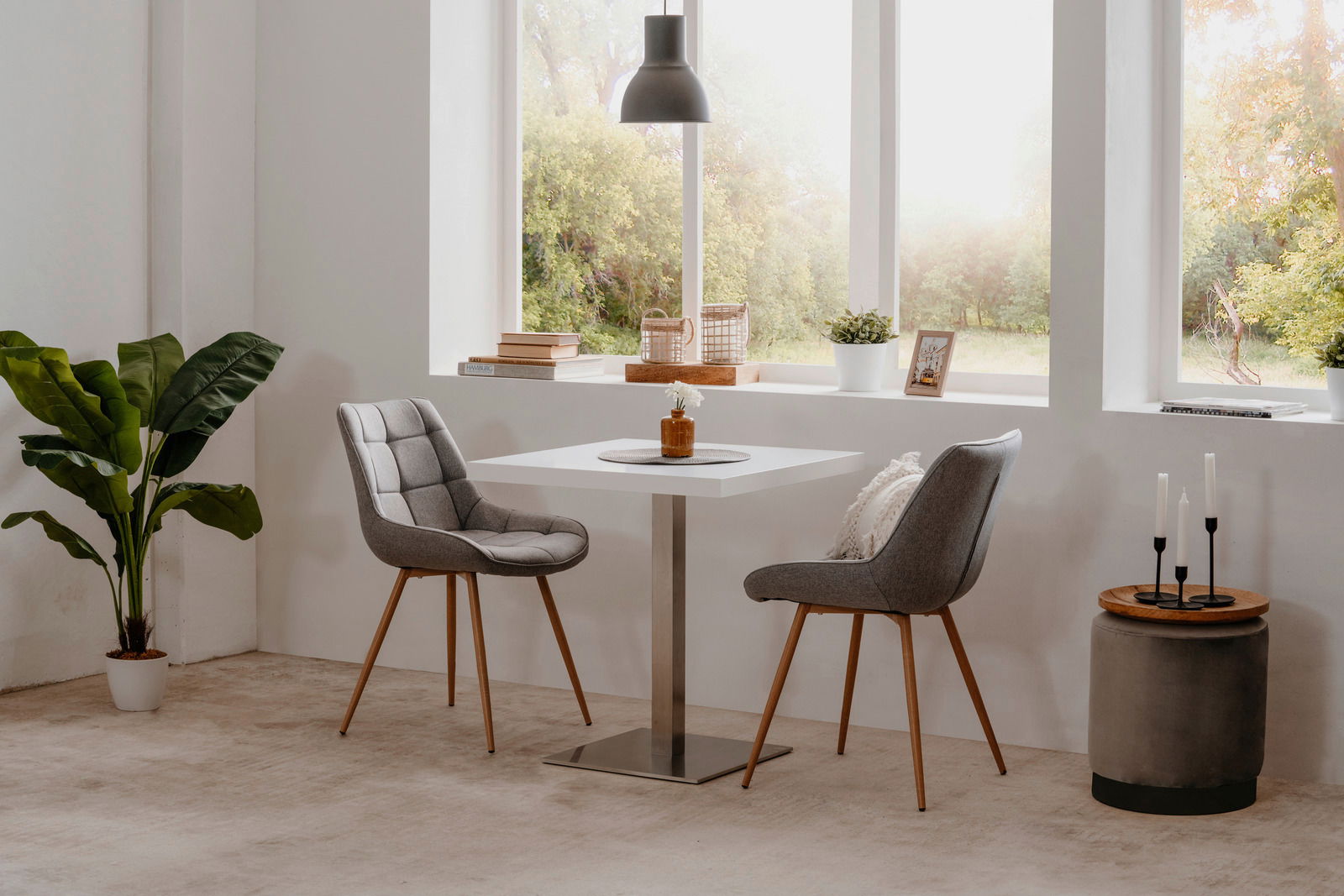 Moderner Esstisch mit quadratischer weißer Tischplatte und Metallfuß, umgeben von zwei grauen Polsterstühlen mit Holzbeinen, in einem hellen Raum mit großen Fenstern und Blick ins Grüne, aus seitlicher Perspektive.