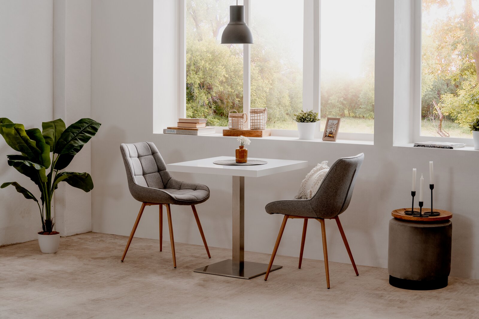 Moderner Esstisch mit quadratischer weißer Tischplatte und Metallfuß, umgeben von zwei grauen Polsterstühlen mit Holzbeinen, in einem hellen Raum mit großen Fenstern und Blick ins Grüne, aus seitlicher Perspektive.