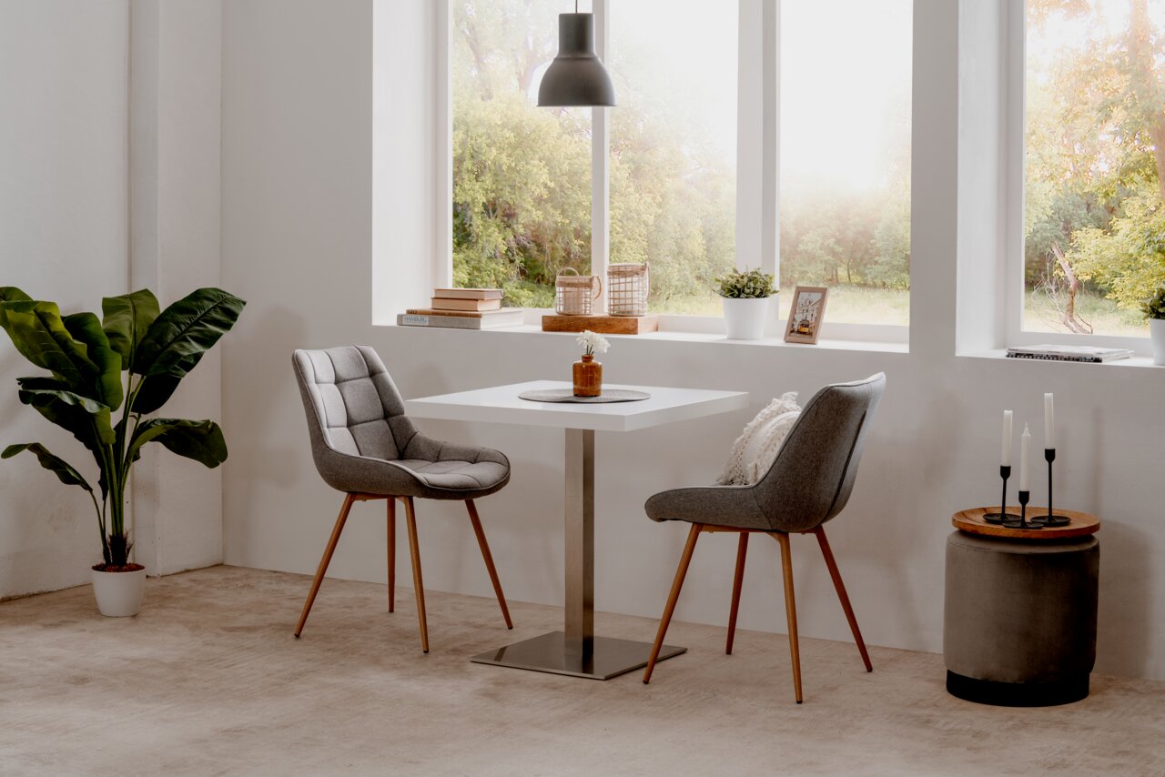 Moderner Esstisch mit quadratischer weißer Tischplatte und Metallfuß, umgeben von zwei grauen Polsterstühlen mit Holzbeinen, in einem hellen Raum mit großen Fenstern und Blick ins Grüne, aus seitlicher Perspektive.