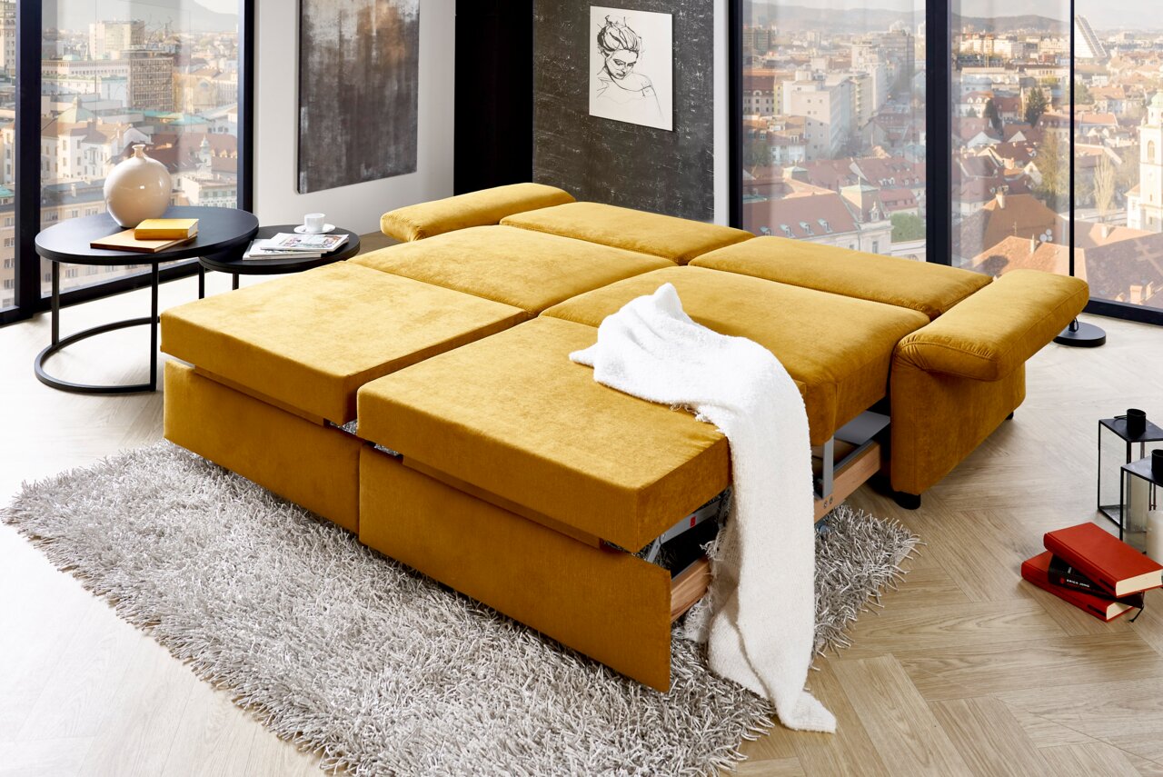 Gelbes Funktionssofa in ausgeklappter Position, seitliche Perspektive, mit weißer Decke auf einem grauen Teppich in einem modernen Wohnzimmer mit Stadtblick.