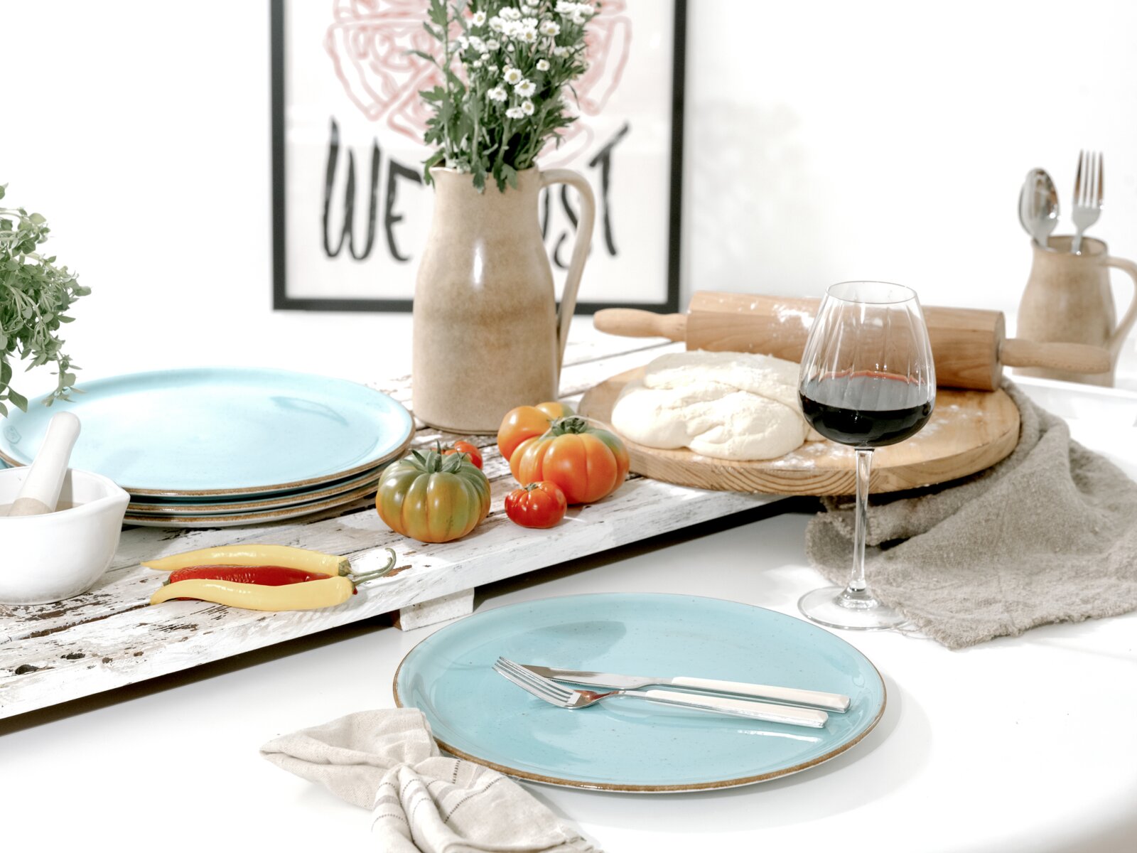 Heller, rustikaler Esstisch mit türkisfarbenen Keramiktellern, Besteck, Rotweinglas, Teig auf Holzbrett, Tomaten und Keramikvase mit Blumen, aufgenommen aus leicht erhöhter Frontperspektive.