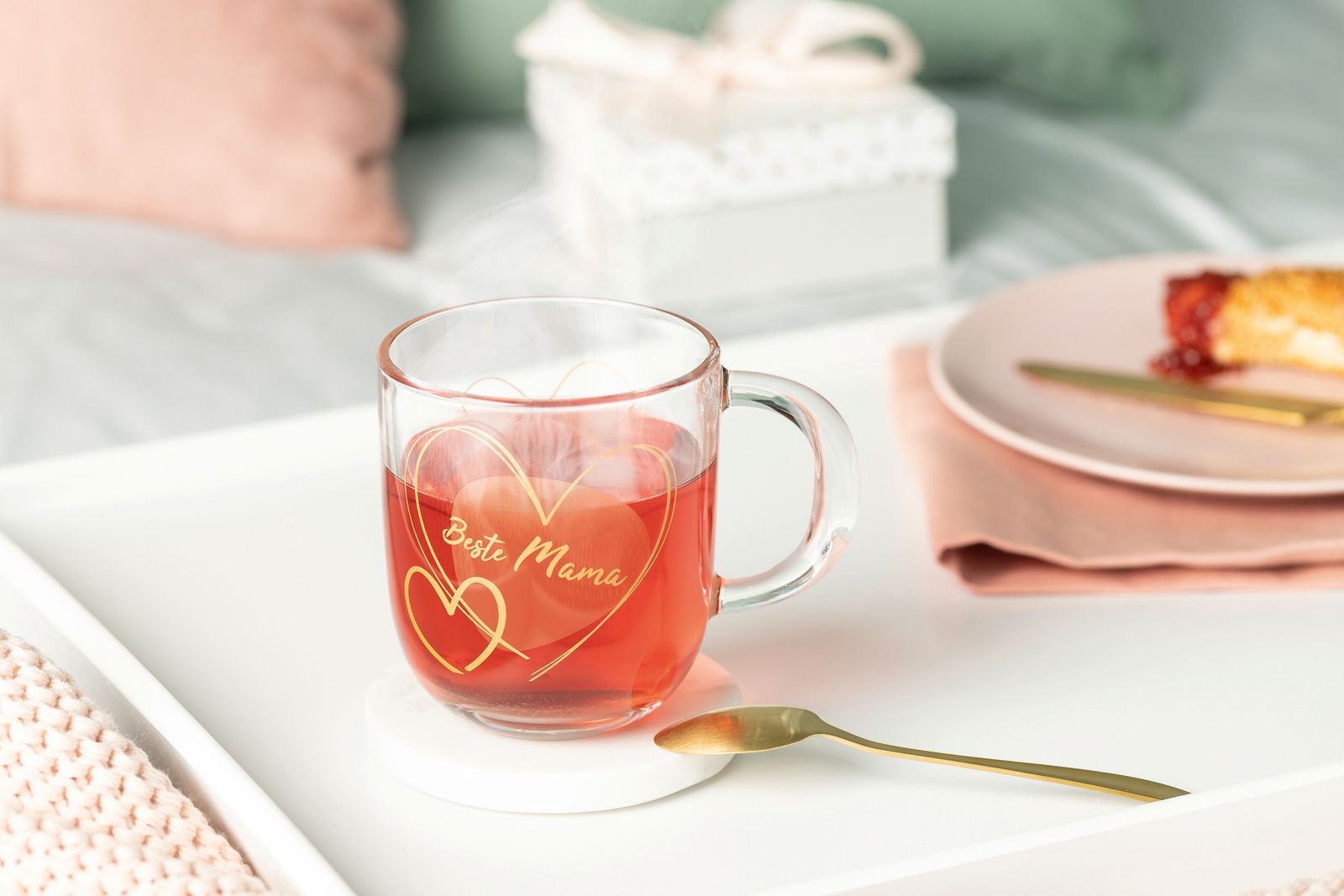 Leonardo Geschenktasse Beste Mama EMOZIONE Glas-Tasse mit der Aufschrift 'Beste Mama' und Herzmotiven, gefüllt mit heißem Tee, auf einem weißen Tablett mit Löffel, aus seitlicher Perspektive fotografiert.