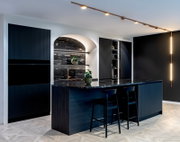 Moderne Designküche mit dunkler Kücheninsel und schwarzer Naturstein-Arbeitsplatte, zwei Barhockern, integrierter Beleuchtung und offenem Regal, aufgenommen aus leicht schräger Frontansicht im Weitwinkel.
