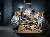 Eine Gruppe von Menschen sitzt um einen Tisch und genießt ein gemeinsames Essen mit der Lumero Gourmet Station 3-in-1, die zentral auf dem Tisch platziert ist. Die Perspektive zeigt das Gerät von vorne, während die Personen fröhlich miteinander interagier