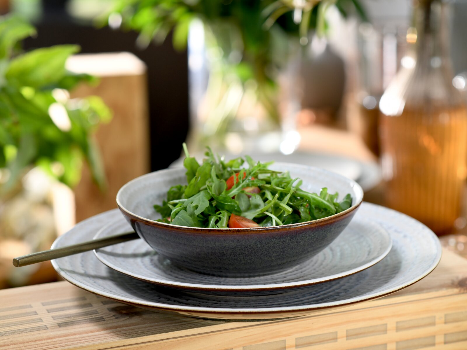 Graue Keramik-Schale mit Rucola- und Tomatensalat auf passendem Teller-Set, seitliche Perspektive auf dem Tisch mit unscharfem Küchenhintergrund