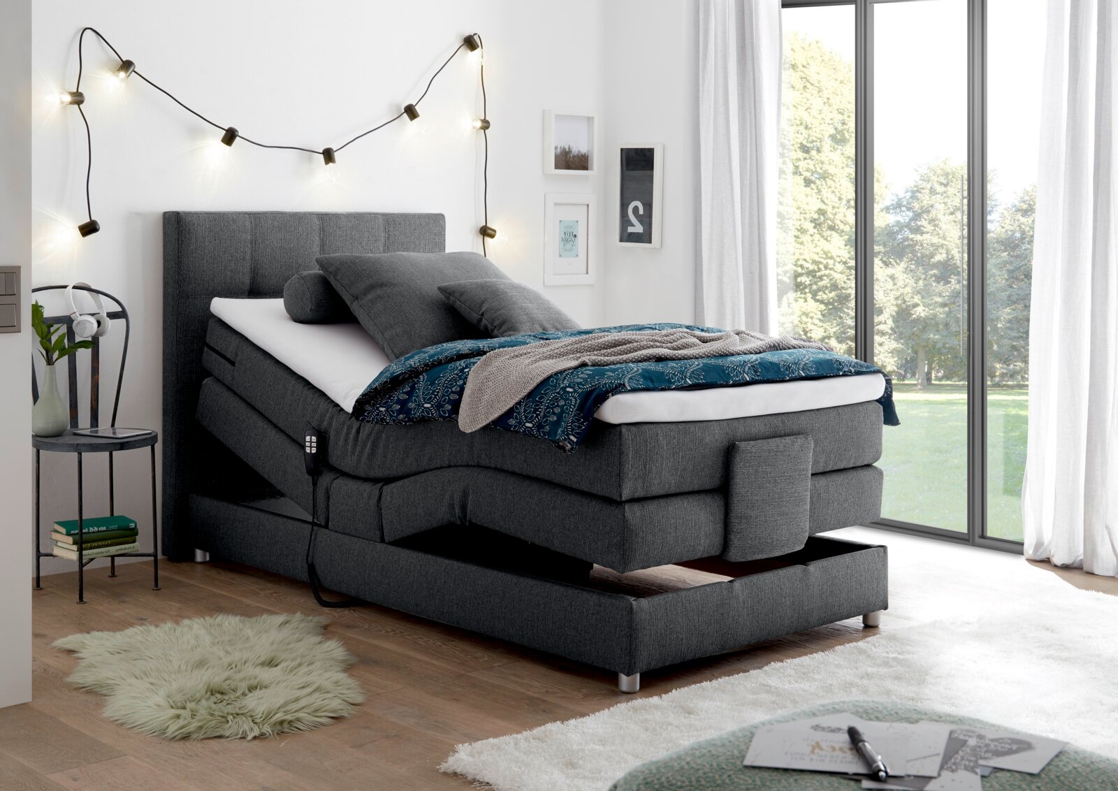 Graues Boxspringbett mit verstellbarem Kopfteil, dekoriert mit Kissen und Decke, in einem modernen Schlafzimmer mit großen Fenstern, aus seitlicher Perspektive.